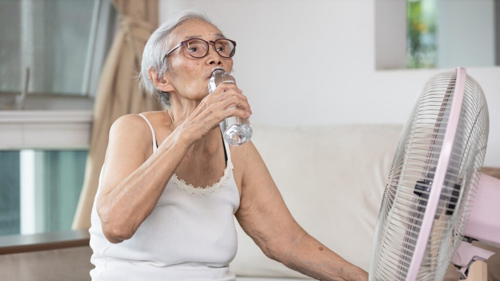 Older adult drinking water while staying cool with a fan during hot weather.